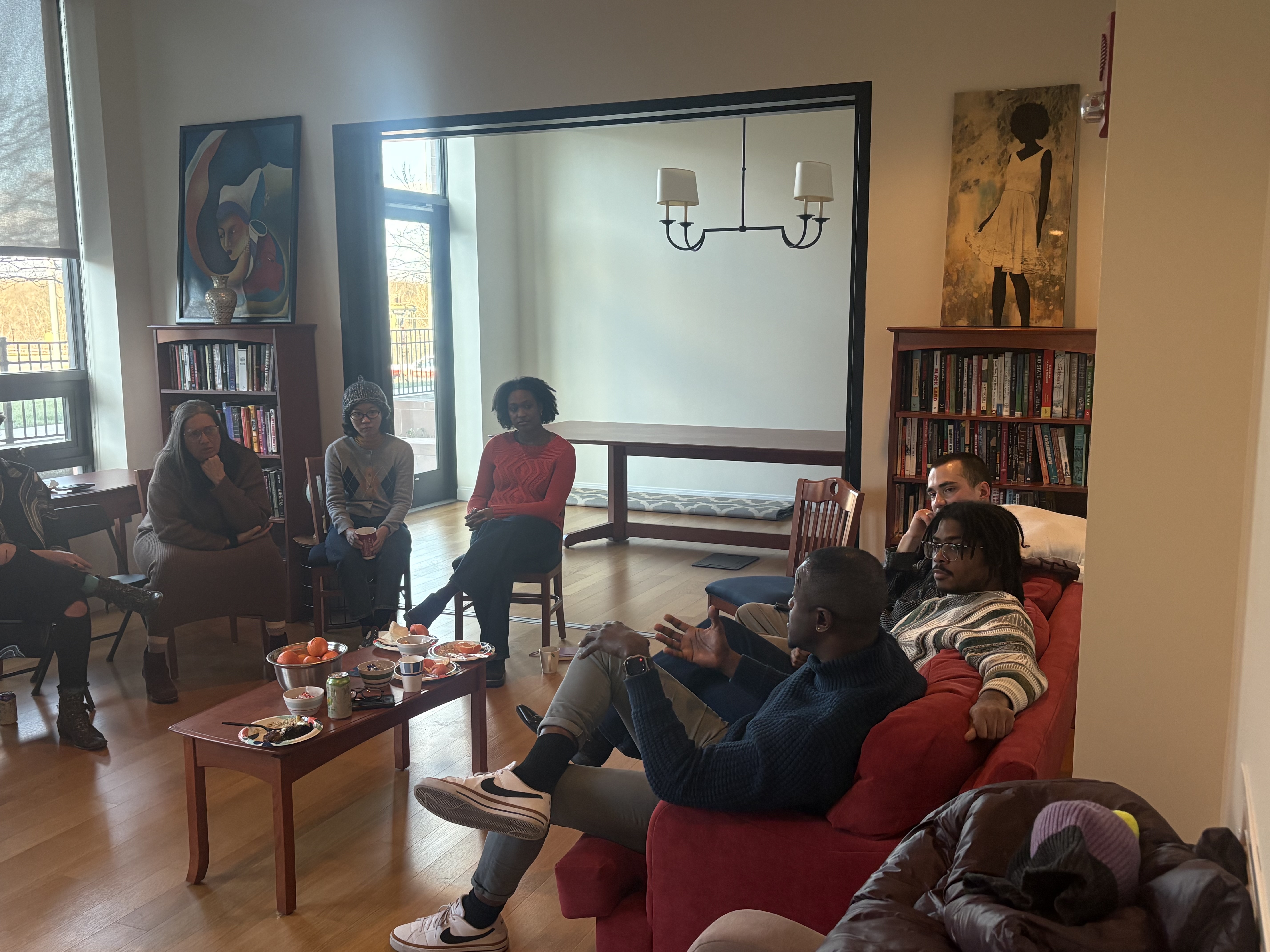 A group of people sits in a circle in a bright, book-lined meeting space during a discussion. Several participants are seated on wooden chairs while two people sit on a red couch in the foreground, with one person speaking and gesturing as others listen attentively. A small coffee table in the center holds fruit, snacks, drinks, and plates. Large windows provide natural light, and bookshelves and artwork decorate the room, creating a warm, welcoming atmosphere for conversation or a seminar.
